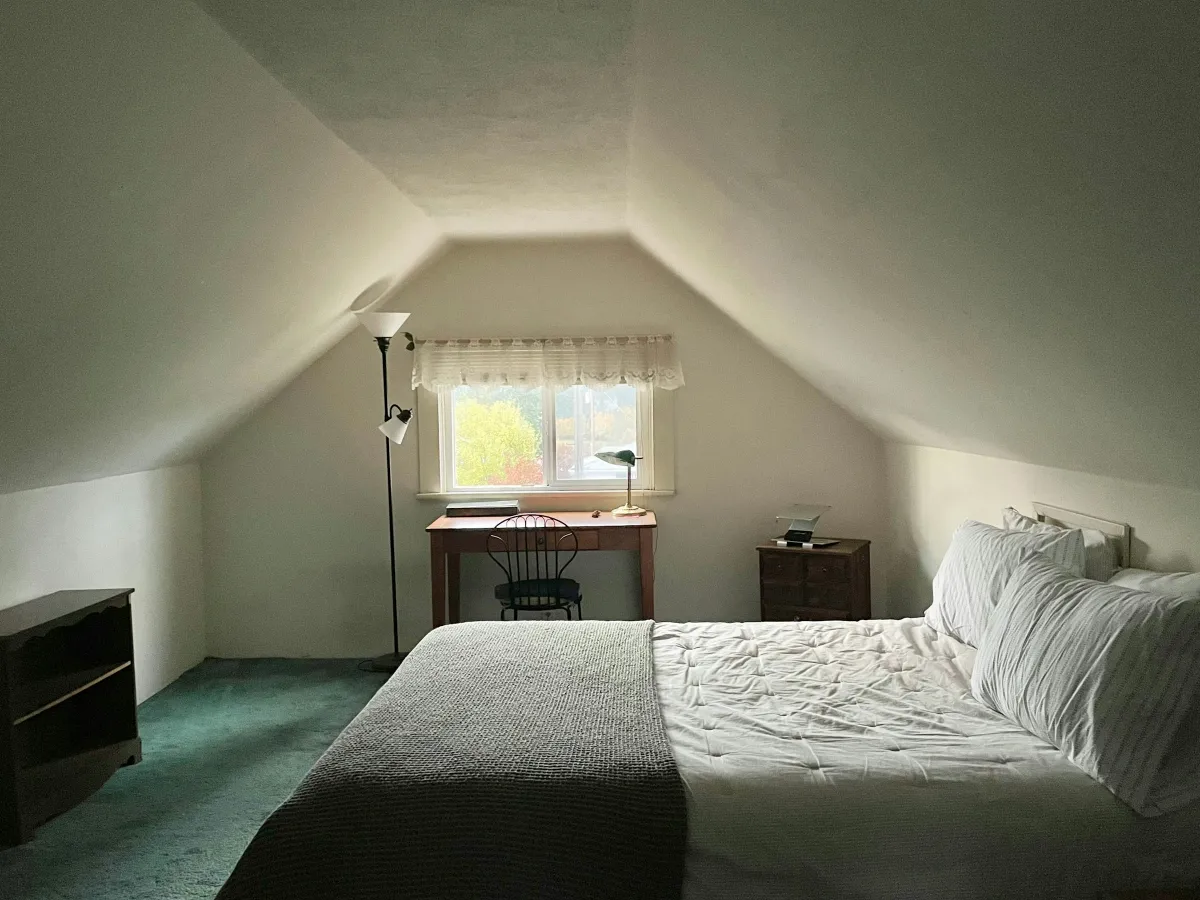 Suite tầng áp mái trần dốc: giường lớn ga trắng, bàn làm việc dưới cửa sổ mái và đèn đứng